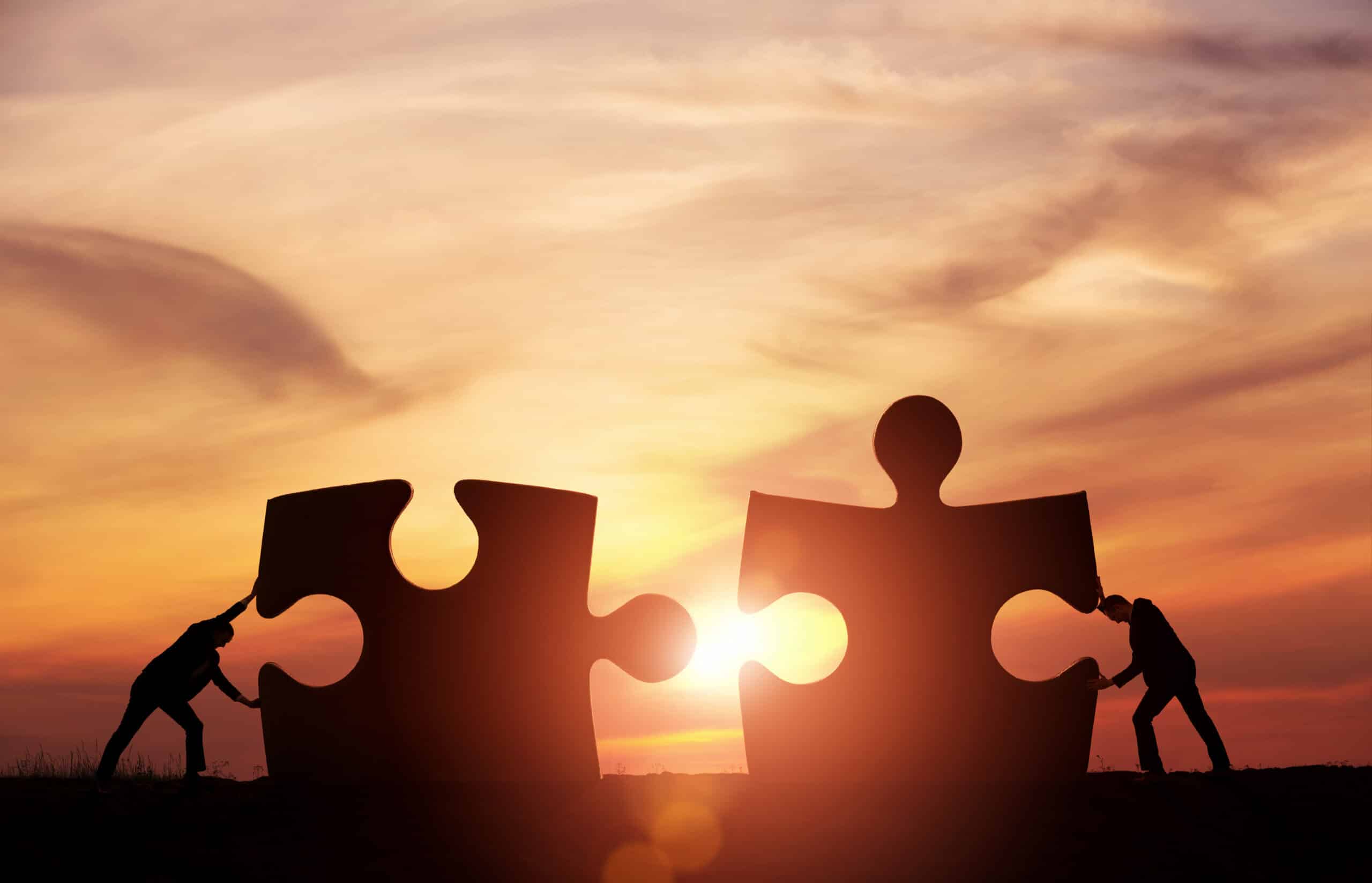 An image of two men pushing together two large puzzle pieces. The sun is setting in the background and the overall image has a dark orange hue.