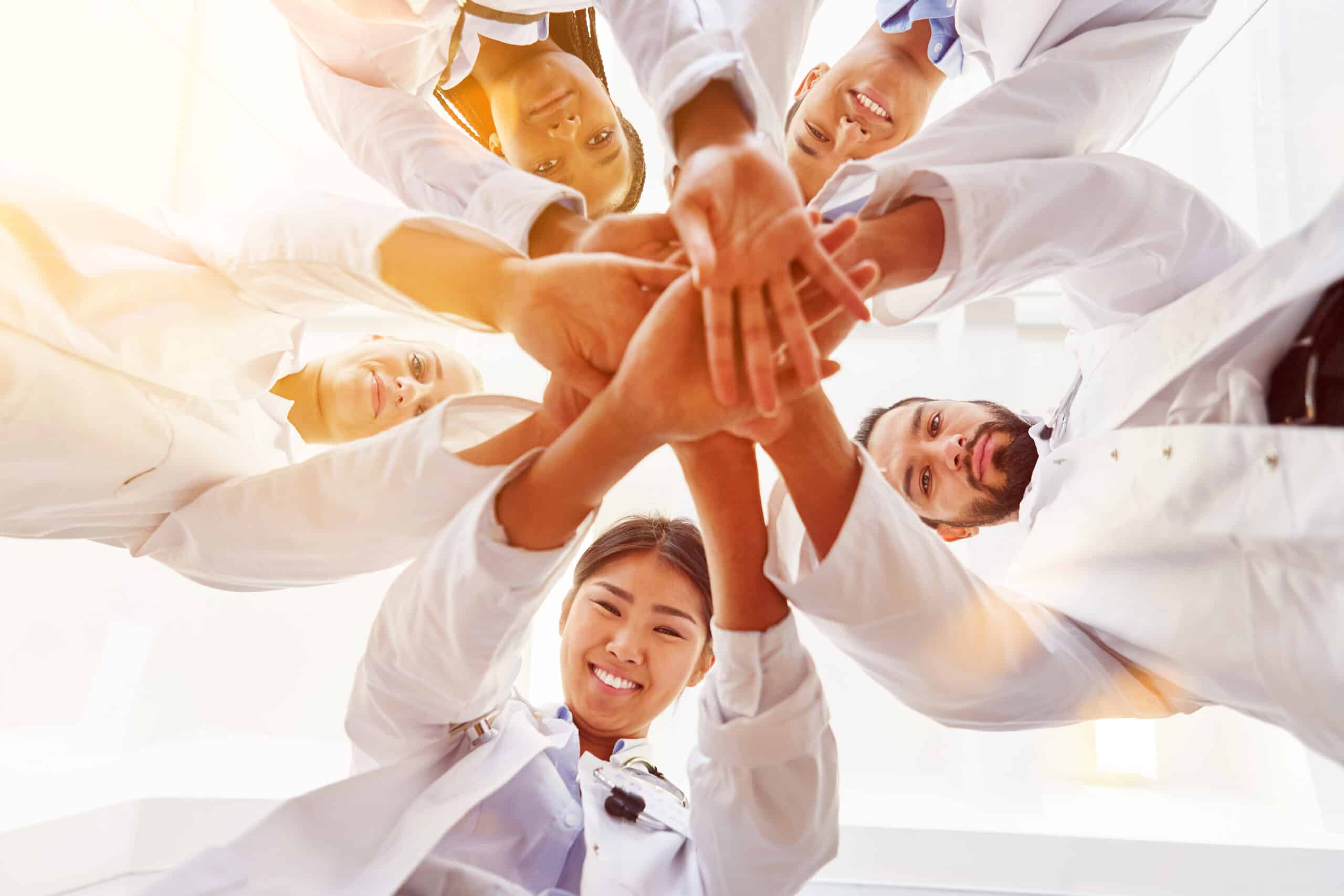 A photo of 5 scientists wearing lab coats and putting their hands in a huddle. They are all smiling down at the camera.